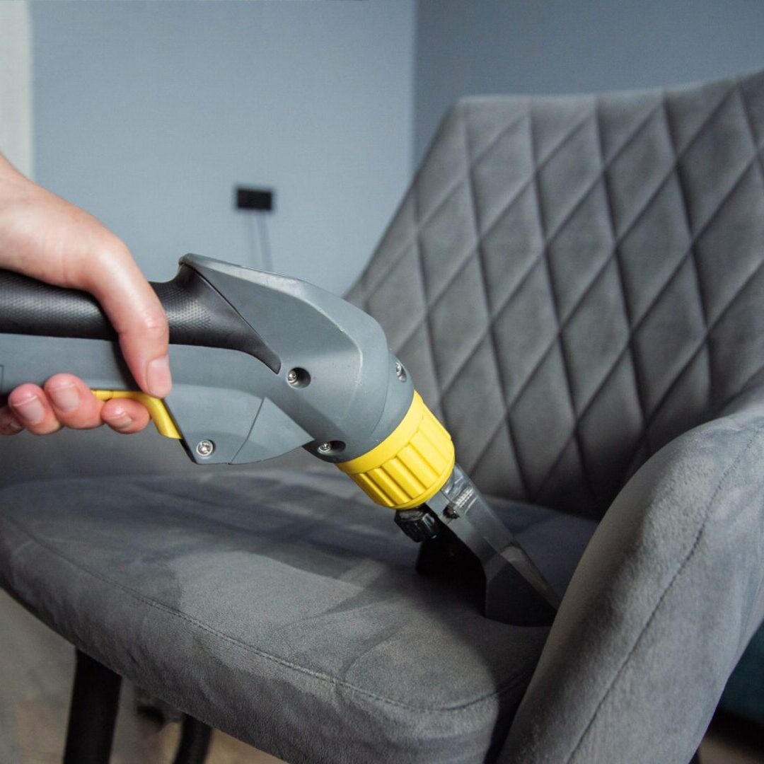 The hands of a girl with a washing vacuum cleaner clean and wash the gray upholstery of a soft chair from dirt. Cleaning services, business, close-up. Copy space for text Vacuuming grey chair with handheld cleaner.